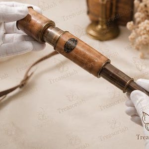 Könnte beinhalten: Ein tragbares Fernrohr aus Messing und Holz mit einem zylindrischen Körper in polierter Ausführung, einem dunklen Metallokular und einer kleinen ovalen Plakette mit eingraviertem Text. Ein dünner brauner Lederriemen ist seitlich befestigt.