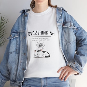 Puede incluir: Camiseta blanca con el texto "OVERTHINKING" y un gráfico de un gato con una burbuja de pensamiento. La burbuja de pensamiento contiene un garabato. El texto debajo de la palabra "OVERTHINKING" dice "BECAUSE MY MIND LOVES PROBLEMS THAT DON'T EXIST."