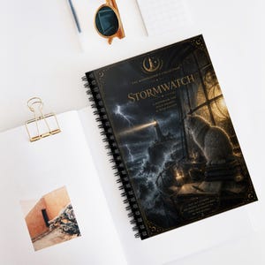 Op de afbeelding: Spiraalgebonden notitieboek met de titel "Stormwatch" met een dramatische illustratie van een vuurtoren tijdens een storm. De omslag toont een kat die uit een raam kijkt. Het notitieboek ligt open op een wit oppervlak met een gouden paperclip en zonnebril.