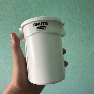 May include: A white cylindrical plastic container featuring the black text BRUTE and the Rubbermaid Commercial Products logo printed on the upper front section. The container has a molded rim with integrated side handles and a smooth finish.