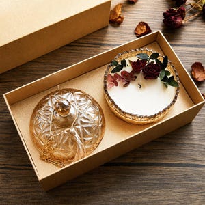 Peut inclure: Une bougie décorative dans un récipient en verre à bordure dorée, ornée de fleurs séchées et de verdure, est présentée dans une boîte dorée. À côté, un couvercle en verre transparent à motifs de diamants avec un gland doré. La bougie est blanche.