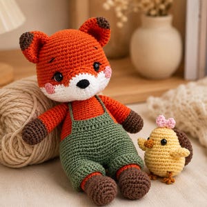 Op de afbeelding: Een gehaakte vossendoll met oranje vacht, bruine accenten en groene tuinbroek zit naast een bol wol. Er is ook een kleine gehaakte gele kuiken met een roze strik. Handgemaakt speelgoed.
