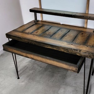 May include: A rustic wooden desk with a black metal frame and a drawer. The desk has a dark wood finish and a distressed look. The desk is supported by hairpin legs.