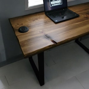 May include: A wooden desk with black metal legs. The desk has a live edge and a natural wood finish. A laptop computer is on the desk with the text "Zombie Woodworks" on the screen. There is a black power outlet on the desk.