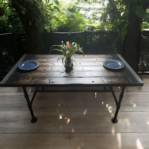 May include: A rustic wooden table with a dark finish and metal legs. Two blue plates and a potted plant with green leaves and a yellow flower sit on the table. The table is on a wooden deck.