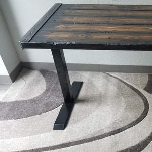 May include: A dark wood desk with a black metal frame. The desk has a rustic look and is made of reclaimed wood. The desk is sitting on a rug with a wavy pattern.