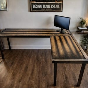 May include: L-shaped wooden desk with a dark stained finish and black metal legs. The desk has a monitor on it and is in a room with a window and a sign that says "DESIGN. BUILD. CREATE."