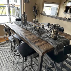 May include: A rustic wooden dining table with a metal frame, adorned with a faux fur runner and decorative candle holders. Black bar stools surround the table, creating a cozy and inviting dining area. The table is 72 inches long.
