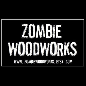 May include: A black background with a white rectangular border containing the words "ZOMBIE WOODWORKS" in a bold, distressed font. Below, in a smaller font, is "WWW. ZOMBIEWOODWORKS. ETSY. COM".