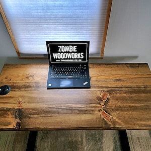 May include: A dark wood desk with a laptop computer on top. The laptop screen displays the text "Zombie Woodworks" and a website address. There is a black electrical outlet on the desk.