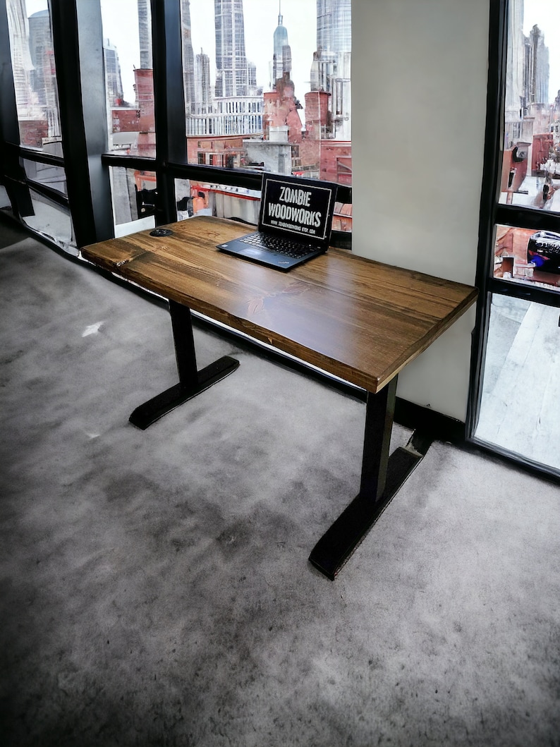 May include: A wooden desk with black metal legs. The desk has a laptop on it with a sticker that says "Zombie Woodworks". The desk is in front of a window with a view of a city skyline.
