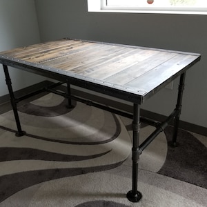 May include: A rustic wooden desk with a black metal frame and legs. The desk has a distressed wood top and a black metal trim around the edge. The desk is sitting on a patterned rug.