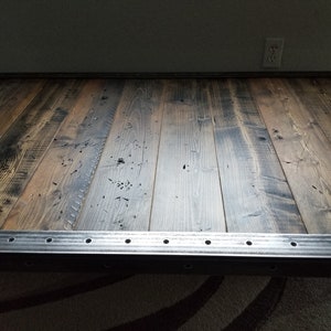 May include: A rustic wooden table top with a metal frame. The wood is dark brown with a distressed finish. The metal frame is silver and has a series of holes along the edge.