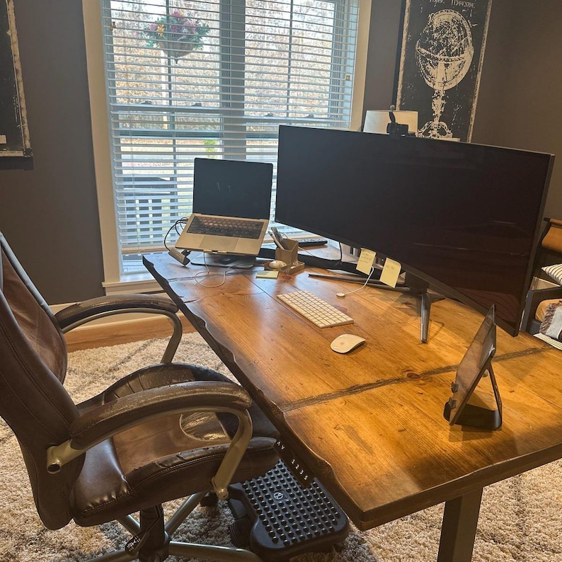 May include: A wooden desk with a laptop, a large monitor, a keyboard, a mouse, and a tablet. A brown leather office chair is in the foreground. The desk is in a home office with a window in the background.