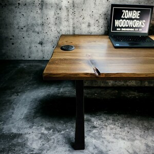 May include: A rustic wooden desk with black metal legs. The desk has a natural wood finish and a small black electrical outlet on the surface. The desk is in front of a concrete wall. The text "ZOMBIE WOODWORKS" is visible on a laptop computer on the desk.
