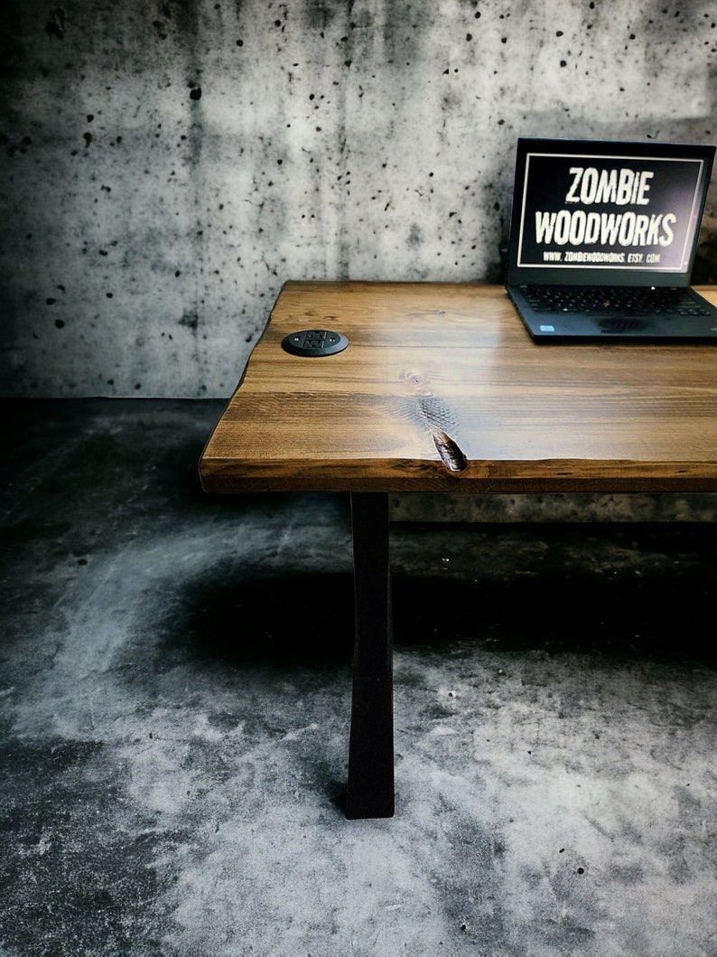 May include: A rustic wooden desk with black metal legs. The desk has a natural wood finish and a small black electrical outlet on the surface. The desk is in front of a concrete wall. The text "ZOMBIE WOODWORKS" is visible on a laptop computer on the desk.