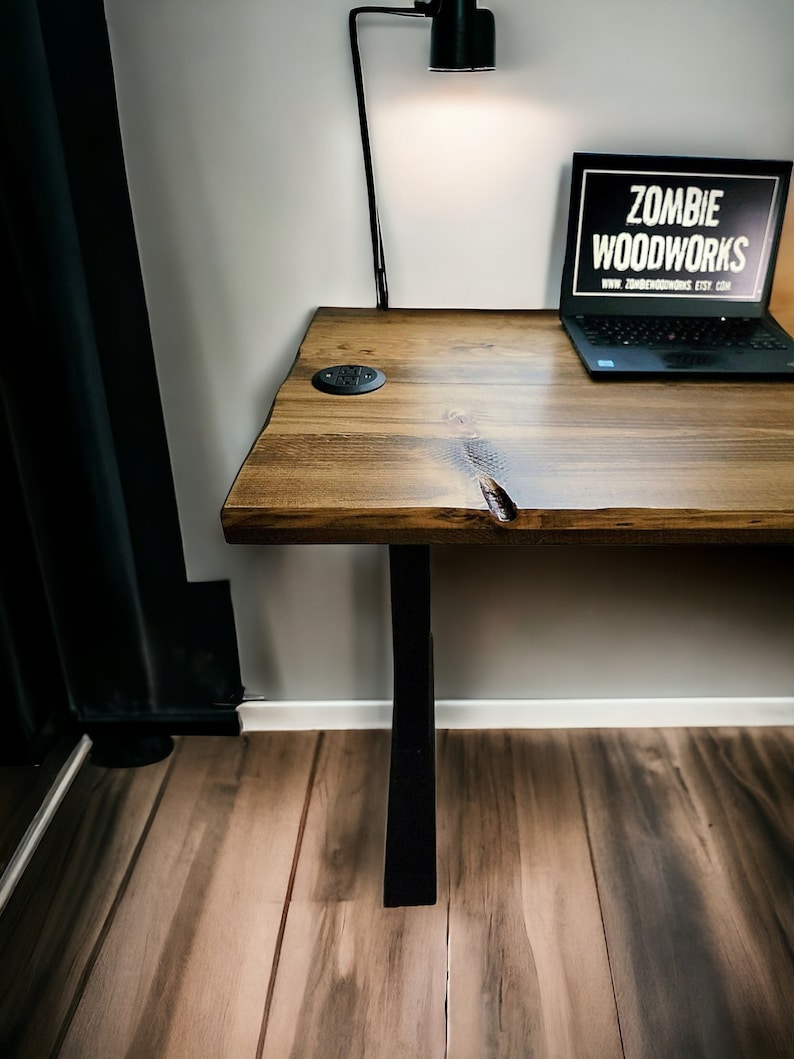 May include: A wooden desk with a black metal leg and a black lamp. The desk has a natural wood finish and a laptop on top of it. The laptop screen displays the text "Zombie Woodworks" and a website address.