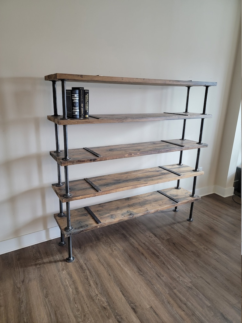 May include: A five-shelf industrial style bookshelf made of reclaimed wood and black metal pipe. The shelves are made of rustic wood with a natural finish. The metal pipes are black and have a matte finish. The bookshelf is standing on a wood floor.