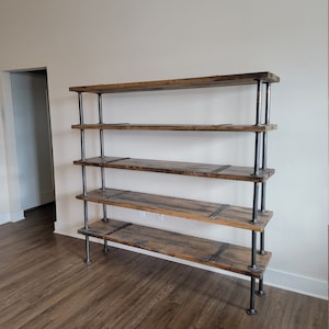 May include: A five-shelf industrial style bookshelf made of reclaimed wood and black metal pipes. The shelves are made of thick, rustic wood planks with a natural finish. The metal pipes are connected with black metal brackets. The bookshelf is standing on a light brown wood floor.