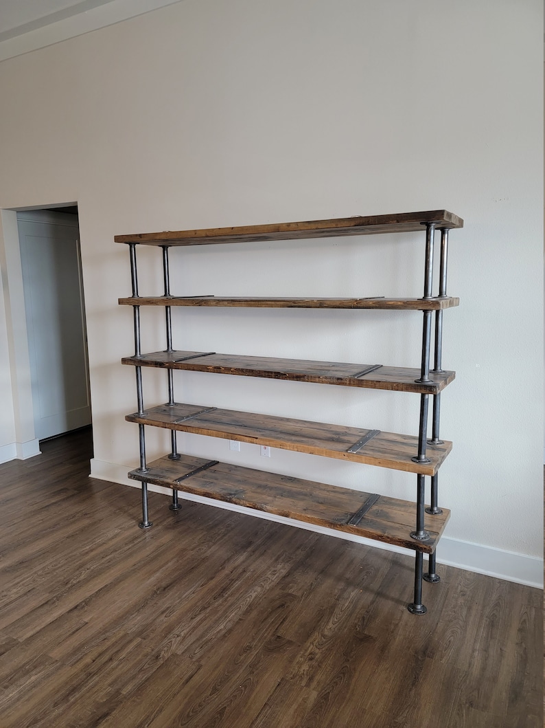 May include: A five-shelf industrial style bookshelf made of reclaimed wood and black metal pipes. The shelves are made of thick, rustic wood planks with a natural finish. The metal pipes are connected with black metal brackets. The bookshelf is standing on a light brown wood floor.