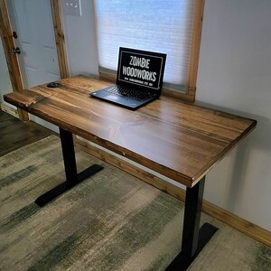May include: A wooden standing desk with black metal legs. The desk has a natural wood finish and is topped with a laptop computer. The laptop screen displays the text "Zombie Woodworks" and a website address.