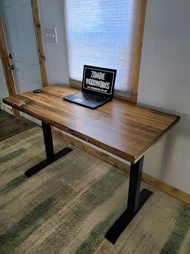May include: A wooden standing desk with black metal legs. The desk has a natural wood finish and is topped with a laptop computer. The laptop screen displays the text "Zombie Woodworks" and a website address.