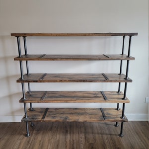 May include: A five-tier industrial-style shelf unit with black metal pipes and reclaimed wood shelves. The shelves are stained a dark brown color and have a rustic look. The unit is freestanding and has a simple, modern design.