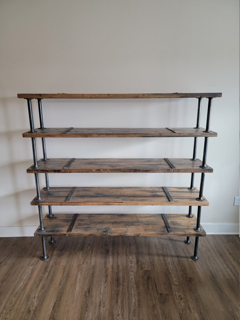 May include: A five-tier industrial-style shelf unit with black metal pipes and reclaimed wood shelves. The shelves are stained a dark brown color and have a rustic look. The unit is freestanding and has a simple, modern design.