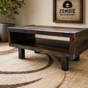 May include: A dark stained wooden coffee table with a lower shelf and metal corner accents. The table is rectangular and sits on four black legs. The sign above the table reads "ZOMBIE WOODWORKS MADE IN THE USA."