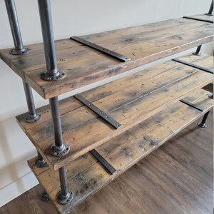 May include: A rustic wooden shelf unit with three shelves supported by black metal pipes. The shelves have a distressed wood finish and are secured with metal brackets.