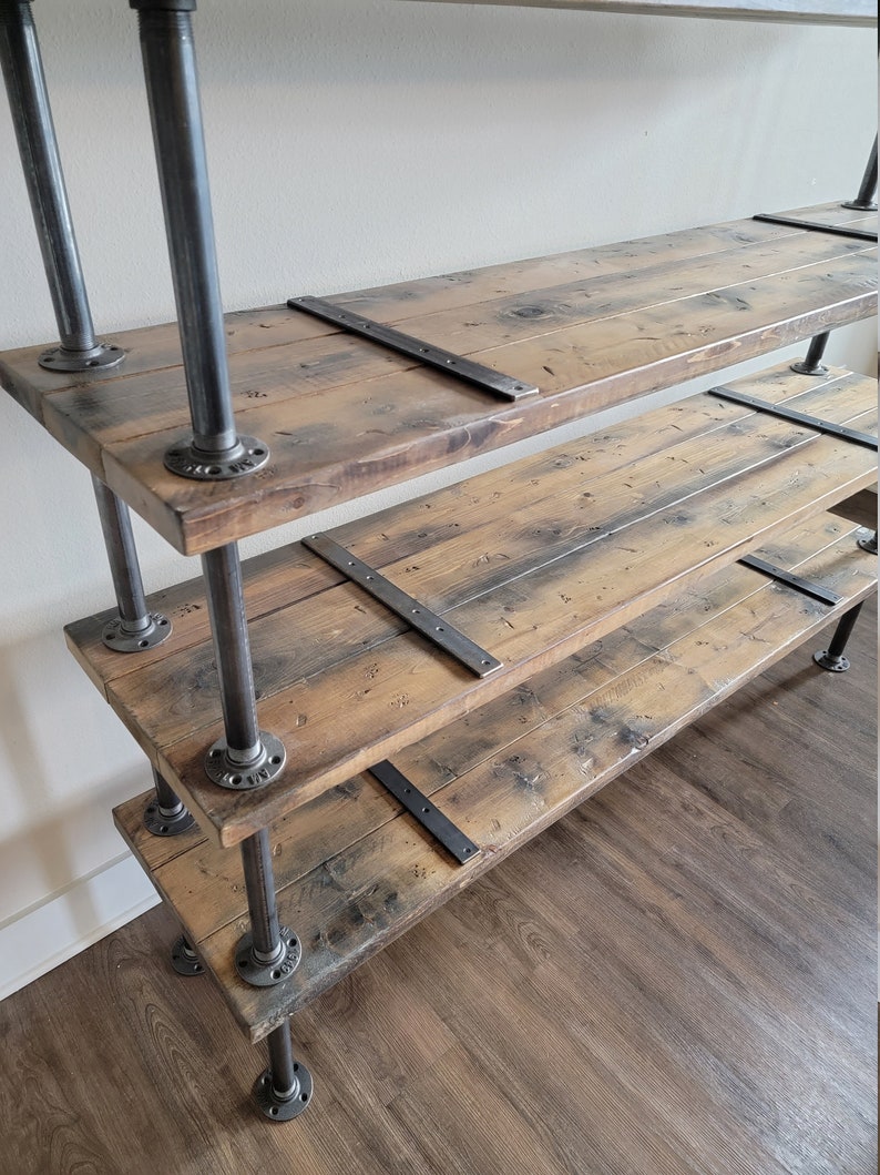 May include: A rustic wooden shelf unit with three shelves supported by black metal pipes. The shelves have a distressed wood finish and are secured with metal brackets.