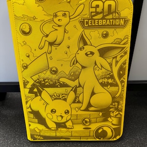 Może przedstawiać: Jaskrawożółty, prostokątny segregator z logo 30th Celebration oraz ilustracjami Mew, Espeona i Pikachu. Okładka przedstawia świąteczną scenę na świeżym powietrzu z balonami i konfetti, utrzymaną w monochromatycznej żółto-złotej kolorystyce.