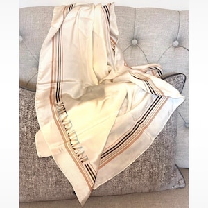 May include: A cream-coloured square scarf featuring a smooth, satin-like finish. The fabric is decorated with thin, parallel brown and black stripes along the border, and the text JOLE VENZIANI is printed in a dark font along one side.