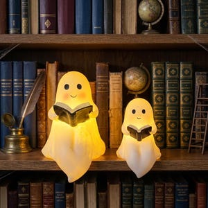 May include: Two glowing, warm-toned ghost-shaped lamps sit on a wooden bookshelf. Each ghost figure has a simple, smiling face and holds an open black book. The lamps emit a soft, diffused light, contrasting with the surrounding vintage books.