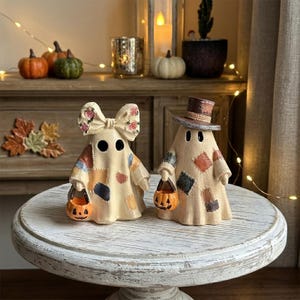 May include: Two ghost figurines with cream-colored bodies featuring patchwork patterns in muted tones. One ghost wears a large bow with a floral print, while the other wears a brown top hat. Each ghost holds a small orange jack-o'-lantern bucket.