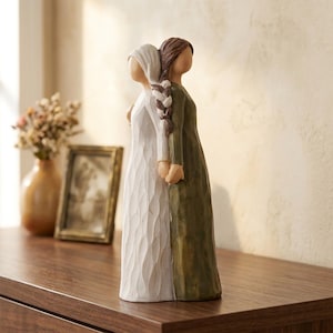Peut inclure: Figurine décorative représentant deux personnages dos à dos, se tenant la main. L'un porte une robe blanche, l'autre une robe verte. Les cheveux sont bruns et tressés. La figurine est sur une surface en bois.