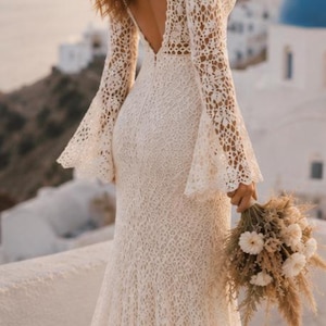 Peut inclure: Une robe longue en dentelle blanche avec des manches longues évasées, un dos nu profond et un corsage ajusté qui s'évase légèrement vers l'ourlet. Le motif floral complexe en dentelle recouvre tout le vêtement, accompagné d'un petit bouquet de fleurs séchées.