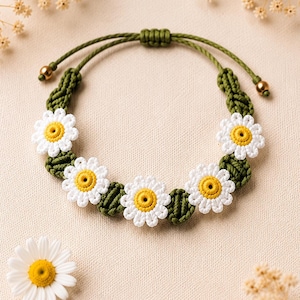 Puede incluir: Una pulsera de macramé hecha a mano con flores de margarita blancas con centros amarillos. Las flores están conectadas por una banda de macramé verde. La pulsera es ajustable y tiene un cordón verde. El fondo es de color beige neutro.
