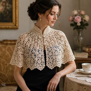 May include: A cream-colored, open-weave crochet capelet with a high collar and button closure. The delicate, lacy design is visible. The capelet is worn over a black dress. The image is set in a room with antique furniture and floral arrangements.