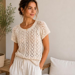 Könnte beinhalten: Cremefarbenes, kurzärmeliges Strickoberteil mit zartem Lochmuster. Das Oberteil hat einen runden Ausschnitt und wird mit einer weißen Hose mit Kordelzug kombiniert. Im Hintergrund steht ein kleiner Olivenbaum in einem gewebten Topf.