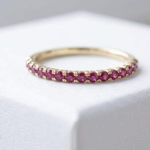 Könnte beinhalten: Ein dünner Goldring mit einer durchgehenden Reihe kleiner, runder, leuchtend pink-roter Rubine in einer gemeinsamen Krappenfassung. Das Metall hat ein poliertes Gelbgold-Finish und die Steine sind in Größe und Farbe einheitlich gehalten.