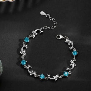 May include: A silver-toned bracelet featuring a series of alternating dolphin-shaped links and round, bright blue faceted stones. The bracelet includes a delicate chain extension with a small tag stamped with the text S925 and a lobster claw clasp.