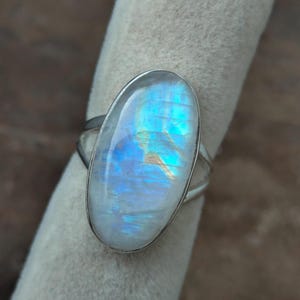 May include: A silver ring featuring a large, oval-shaped moonstone. The moonstone has a translucent, iridescent quality with blue and white hues. The ring's band is a simple, split design.