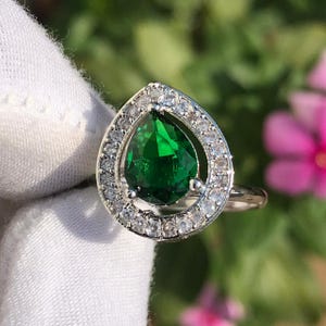 May include: A silver ring featuring a pear-shaped, vibrant green gemstone, surrounded by a halo of smaller, clear stones. The ring is set against a soft, blurred background of green foliage and pink flowers.