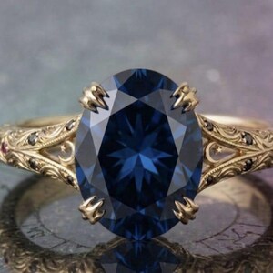 Peut inclure: Une bague en or avec une grande pierre centrale bleu foncé taille ovale fixée par quatre griffes. L'anneau présente des gravures complexes, de petites pierres noires et de minuscules gemmes rouges, avec des motifs d'étoiles et de lunes.
