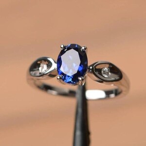 Può includere: Un anello d'argento con una gemma ovale blu scuro come elemento centrale. L'anello ha un design a gambo diviso con piccole pietre di accento chiare su entrambi i lati della pietra principale. L'anello è posto su uno sfondo caldo e neutro.