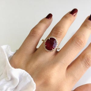 May include: A gold ring featuring a large, oval-cut deep red gemstone at the centre, surrounded by a halo of small, clear, sparkling stones. The band is thin and metallic, providing a delicate frame for the prominent, vibrant red centrepiece.