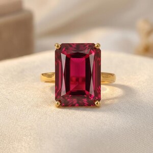 May include: A gold ring featuring a large, rectangular, deep red gemstone. The gemstone is the focal point, set in a gold band with a simple, elegant design. The ring is displayed on a white surface.