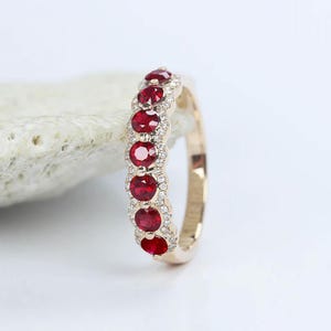 Op de afbeelding: Een gouden ring met een rij van zeven verticaal gezette, rondgeslepen rode robijnen. De robijnen zijn omringd door een delicate halo van kleine, heldere witte diamanten, wat een geschulpte rand langs de ringband creëert. De ring heeft een gepolijste gouden afwerking.