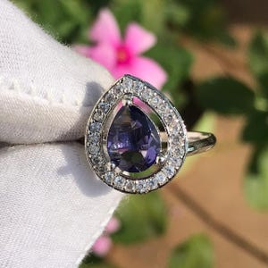 Può includere: Un anello d'argento con una pietra preziosa viola scuro a forma di pera, circondata da un alone di piccole pietre trasparenti. L'anello è posto su uno sfondo bianco morbido, con uno sfondo floreale sfocato.